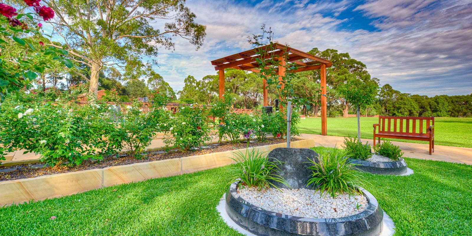 Lush garden scene with green grass, granite circles, rose bushes, and a wooden arbour against a blue sky at Rookwood Crematorium.