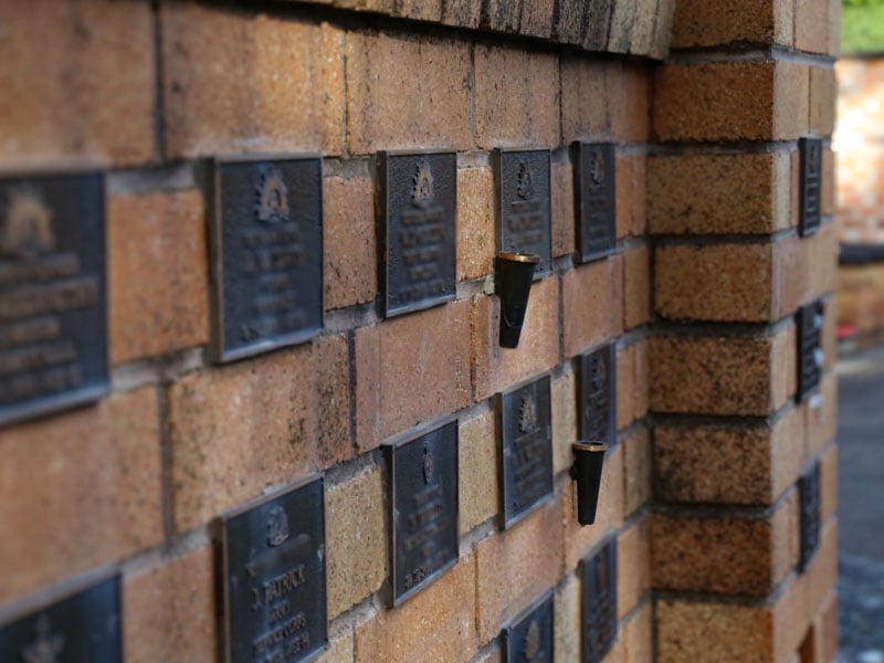 Close-up of a brick wall with evenly spaced bronze plaques, and small metal flower holders at the War Veterans Memorial in Rookwood General Cemetery and Crematorium.