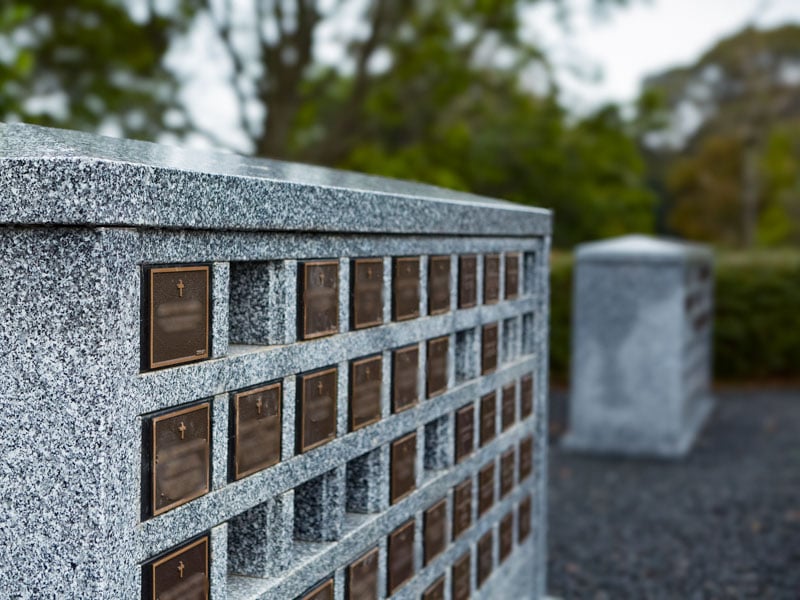 Granite columbarium niche wall with bronze plaques, set in the tranquil Rookwood General Cemetery and Crematorium surrounded by greenery.