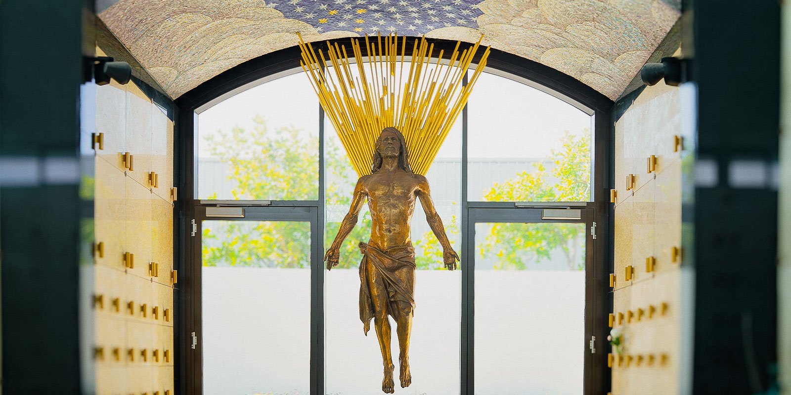 Interior of a memorial space featuring a bronze statue of Christ with radiant golden beams, surrounded by marble niches