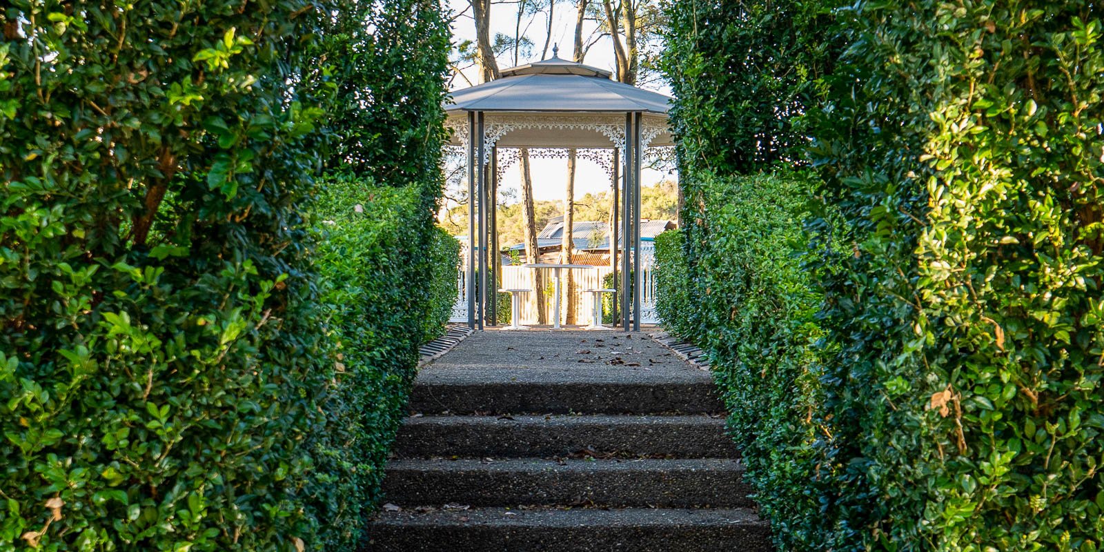 Pathway leading to a white gazebo, framed by lush green hedges in a garden setting