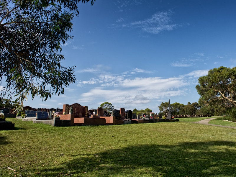 A serene cemetery scene with rows of headstones under a clear, sunny sky. Green grass and trees surround the graves, creating a peaceful atmosphere at Frenchs Forest Bushland Cemetery.