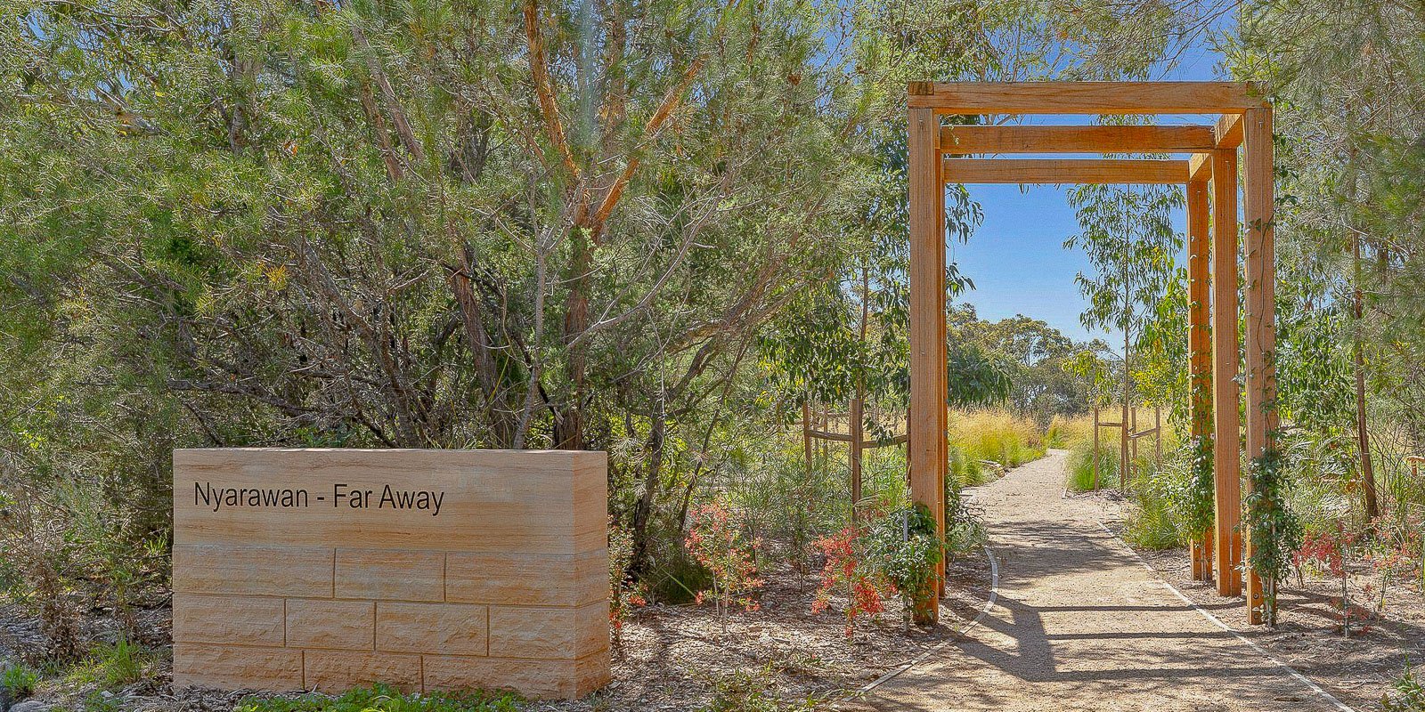 Entrance to a natural burial site with a wooden archway, surrounded by native plants and trees, featuring a sign that reads 'Nyarawan - Far Away
