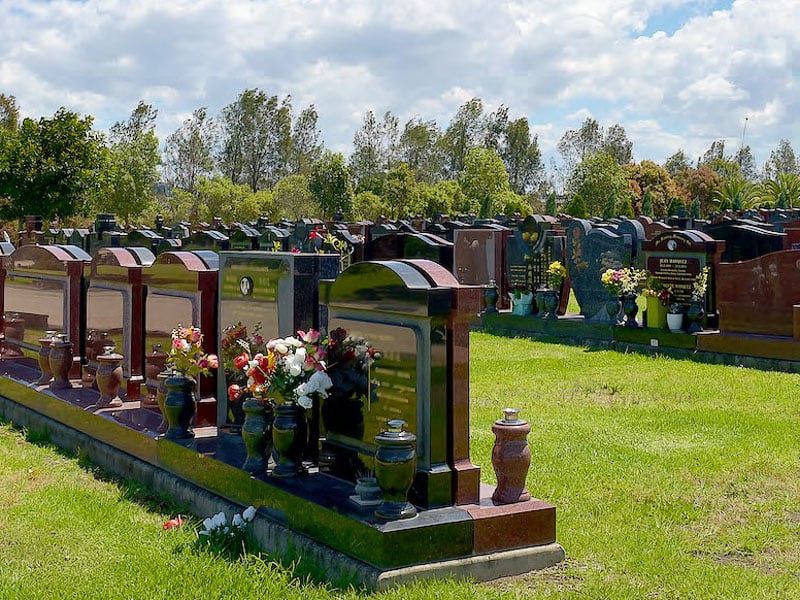 Row of granite headstones in a lawn monumental section, with flower arrangements and urns on green grass.