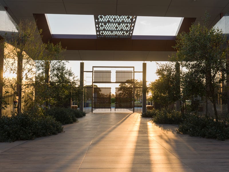 Sunset light casts long shadows in a modern corridor with trees on both sides, leading to a gate at the Holy Mausoleum in Macquarie Park Cemetery.