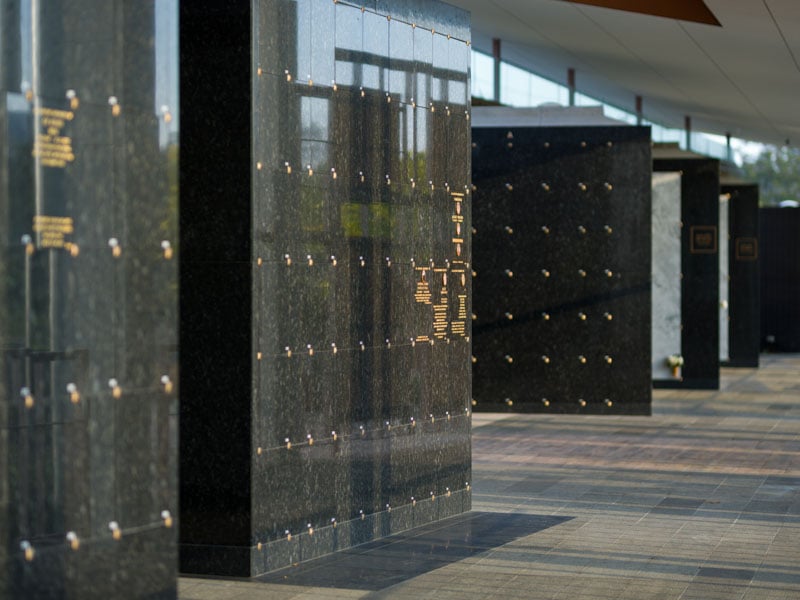 Sunlit columbarium corridor with polished black granite walls, engraved golden plaques, soft reflections, and a calm, respectful ambiance.
