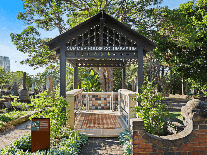 Gazebo labeled "Summer House Columbarium" in a serene cemetery. Lush greenery surrounds, with paths and headstones visible under a clear blue sky.