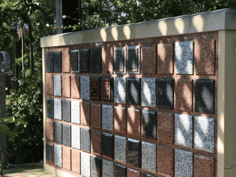 A sunlit Summer House columbarium wall with bronze-colored and gray granite plaques. Shadows from nearby trees fall across the wall, creating a serene atmosphere.