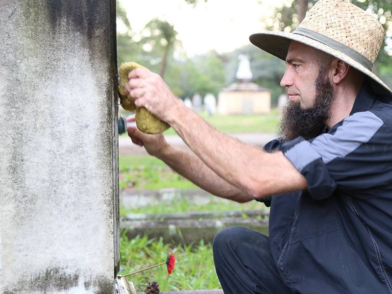 A man in a straw hat cleans a weathered gravestone with a sponge in a cemetery. He is focused and kneeling on the grass.