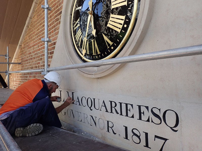 A worker in an orange and blue uniform and white helmet carefully restores letters on a historic building under scaffolding. A large clock with gold Roman numerals is above, set on a brick wall.