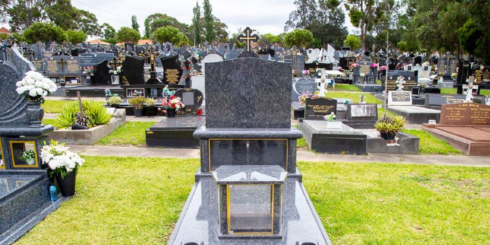 Headstone in Rookwood General Cemetery and Crematorium surrounded by other graves, featuring a granite full monument with a cross and floral arrangements.