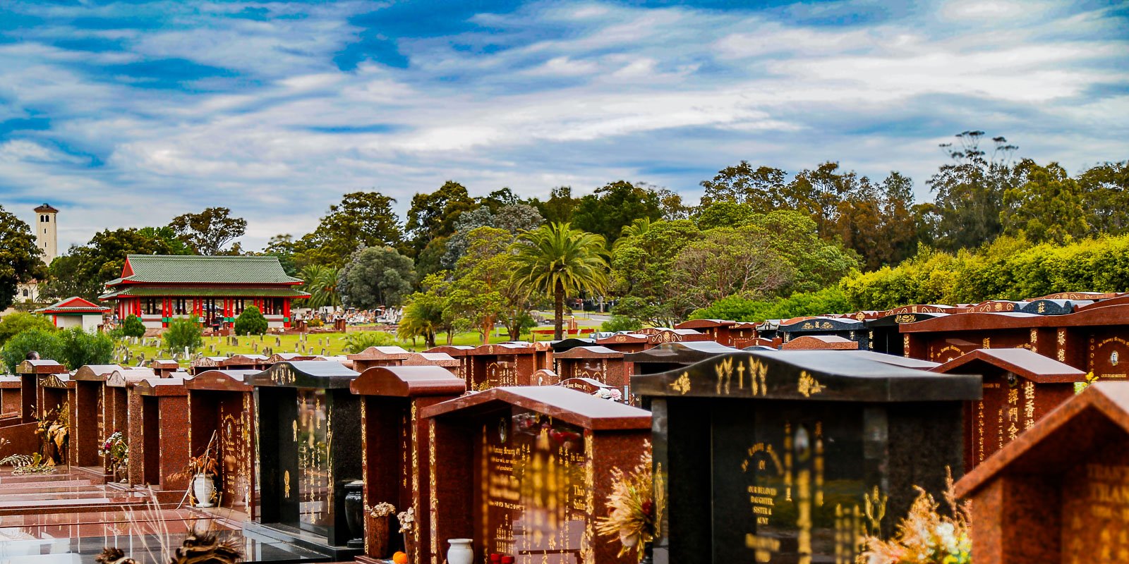 Rookwood General Cemetery and Crematorium with rows of polished headstones and a building with red pillars and green roof in the background, surrounded by lush trees under a cloudy sky.