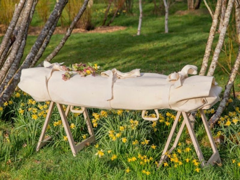 Image of a shrouded body outside surrounded by yellow wildflowers. The shroud is cotton and has flowers placed ontop.