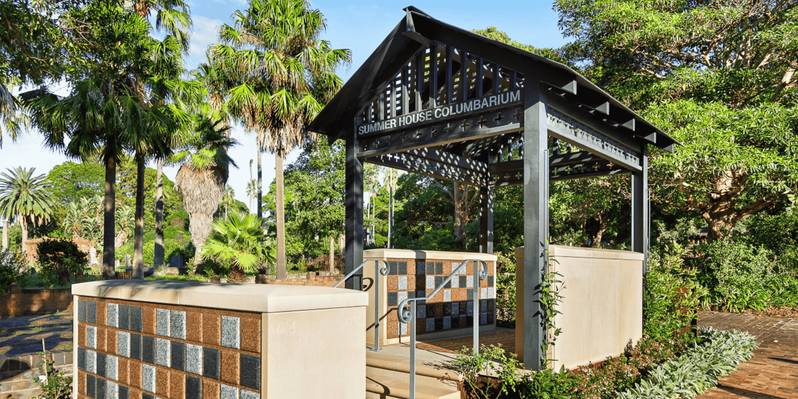An outdoor columbarium named "Summer House" with a tiled wall, set amidst lush greenery and tall palm trees under a clear blue sky, creating a serene atmosphere.
