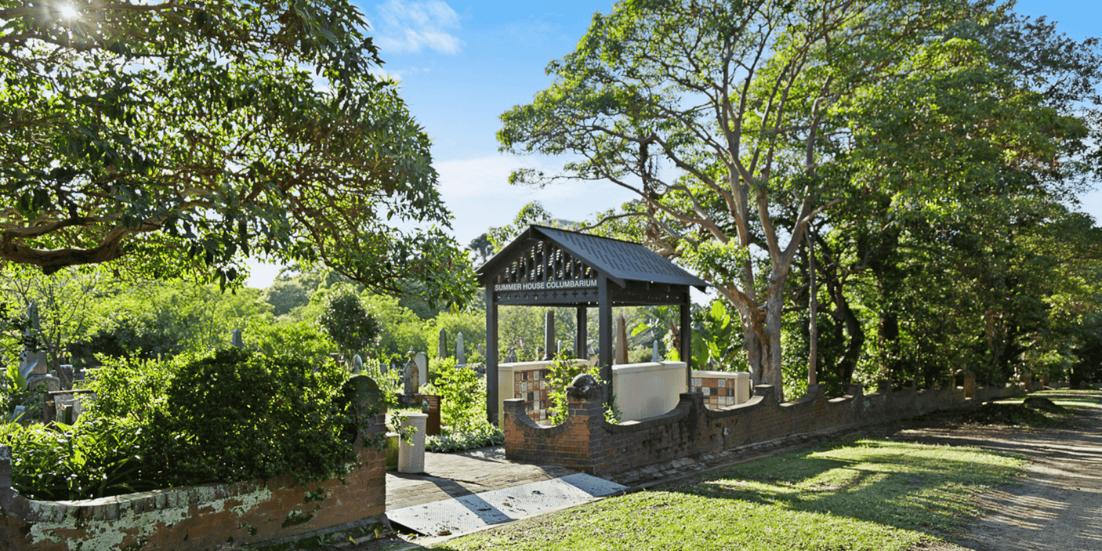 A serene garden with a columbarium entrance under a gabled roof. Surrounded by lush green trees, the sunlight creates a peaceful atmosphere.