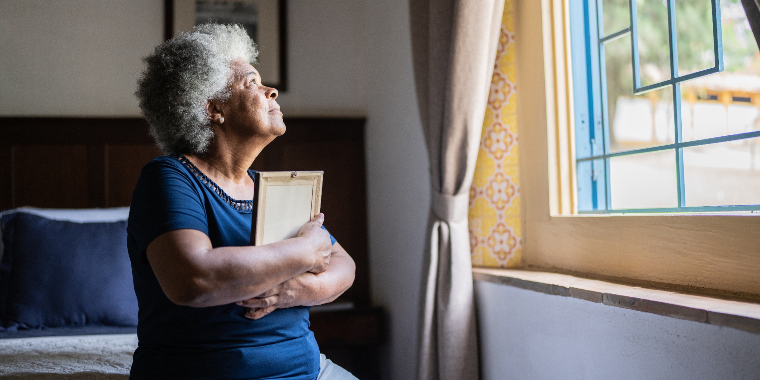 Elderly woman with gray hair gazes out a sunny window, holding a picture frame. Her expression is thoughtful and serene. Room has soft, warm lighting.