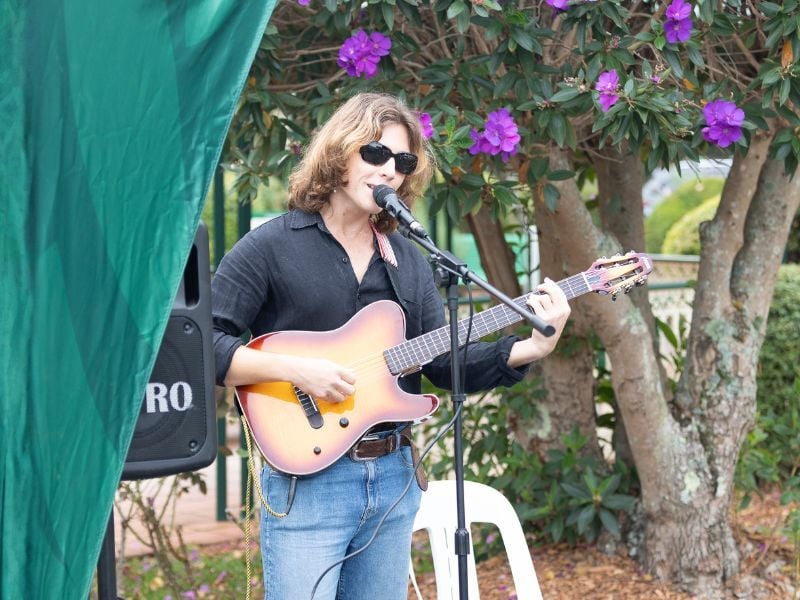A person in sunglasses plays an electric guitar and sings into a microphone outdoors. They stand near a tree with vibrant purple blossoms, creating a relaxed, joyful ambiance.