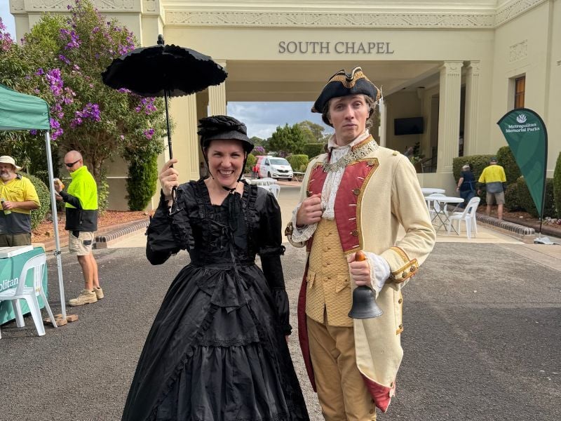 A cheerful woman in a black Victorian dress and hat with a parasol stands beside a man in colonial attire holding a bell, posing outside a chapel.