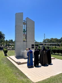 Four clergy in robes stand solemnly before a tall, gray monument on a sunny day, with a vibrant stained glass element. The setting is a landscaped memorial garden.