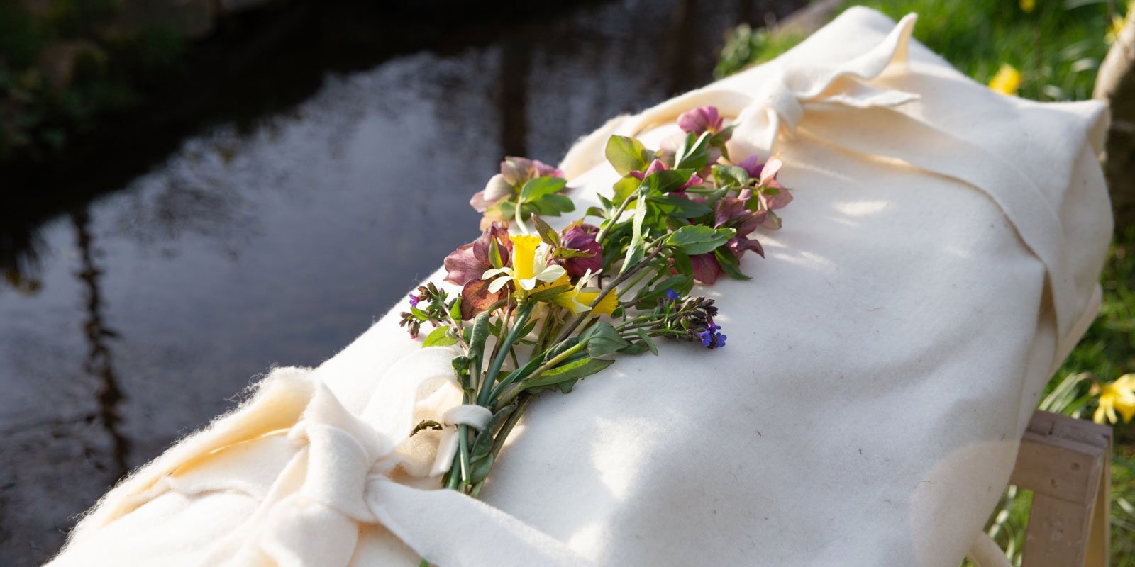 A shrouded body rests on a wooden stand by a tranquil river. A bouquet of colourful flowers adorns the natural fibre cloth.