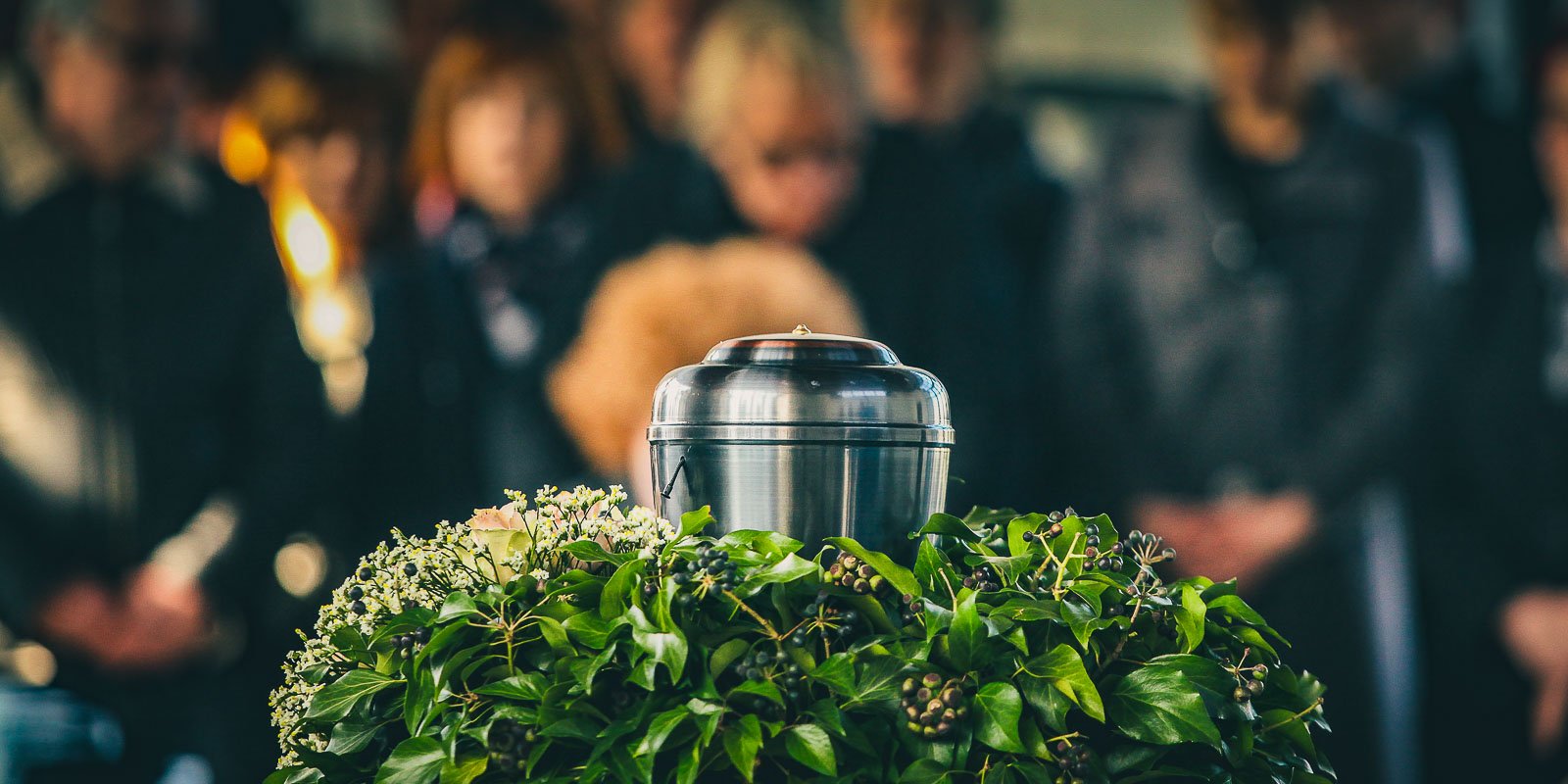 A silver urn resting on a lush arrangement of green leaves and flowers. In the blurred background, somberly dressed people are gathered for a funeral service.