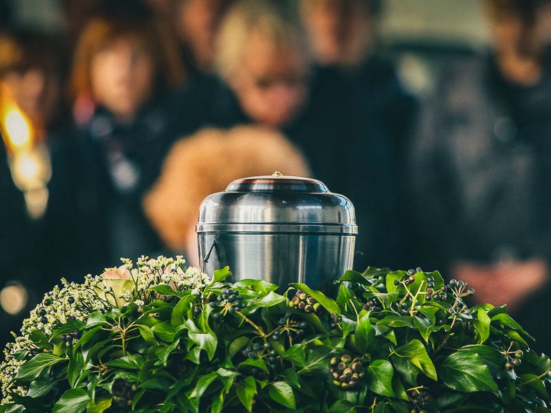 A silver urn resting on a lush arrangement of green leaves and flowers. In the blurred background, somberly dressed people are gathered for a funeral service.