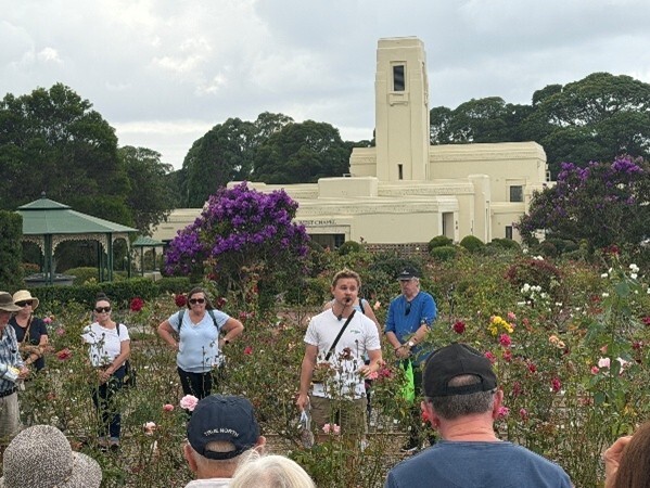 A group of people stand in a vibrant rose garden, listening to a speaker. A historic white art deco building and gazebo are visible in the background.