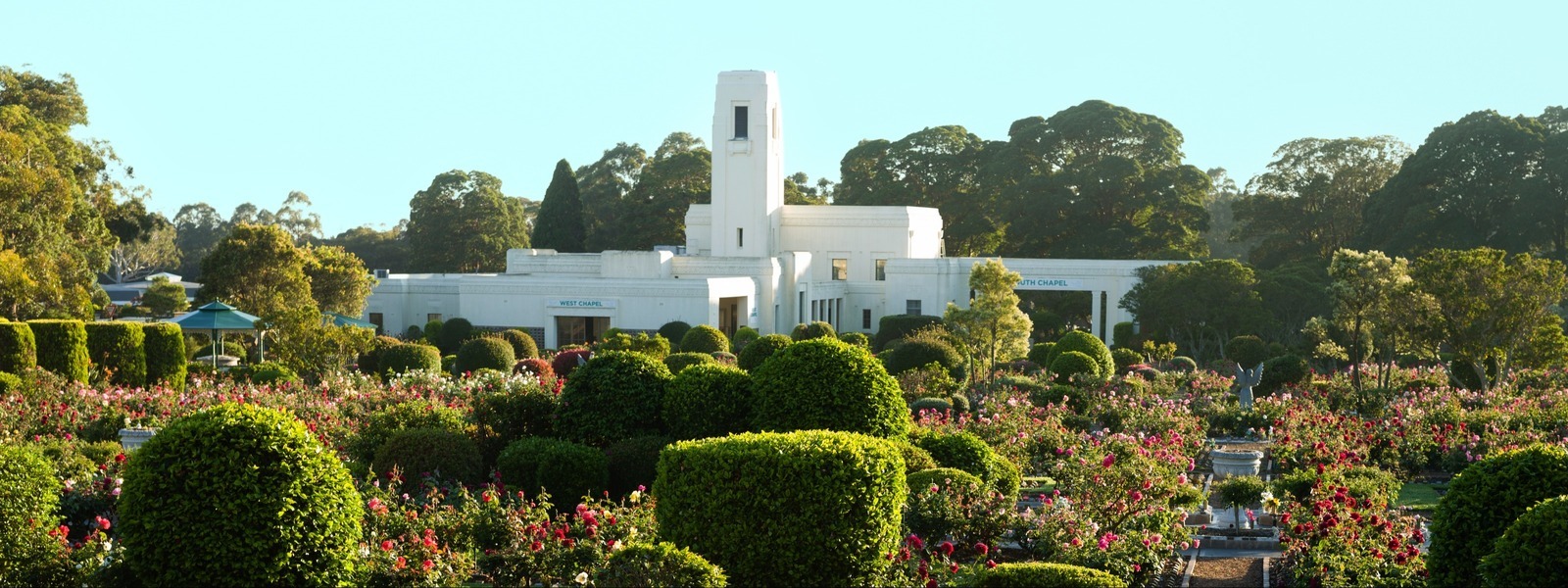 A historic chapel at Woronora Memorial Park, surrounded by well-manicured bushes and flowering plants. The sky is clear and blue.