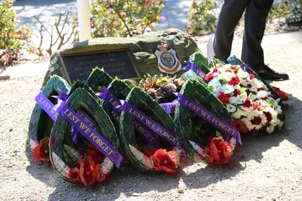 Wreaths with red poppies and "Lest We Forget" ribbons are placed by a memorial plaque on a stone. The tone is solemn and respectful.
