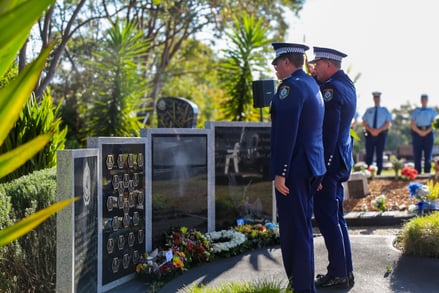 Uniformed police officers stand solemnly before a memorial adorned with plaques and floral tributes, conveying respect and remembrance.