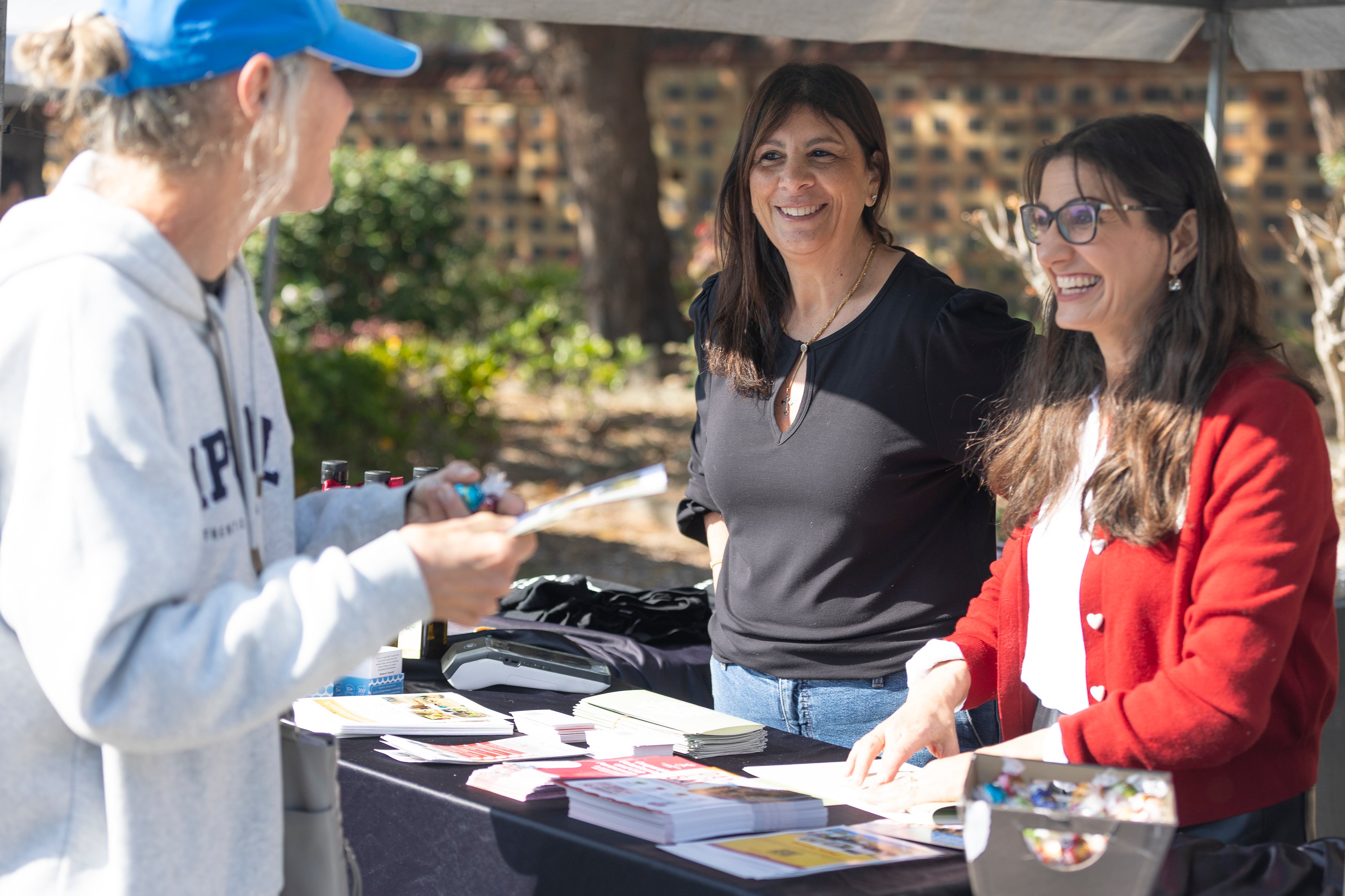 Two smiling women staff a sunny outdoor information table as a visitor in a blue cap points toward brochures, creating a friendly community-event atmosphere.