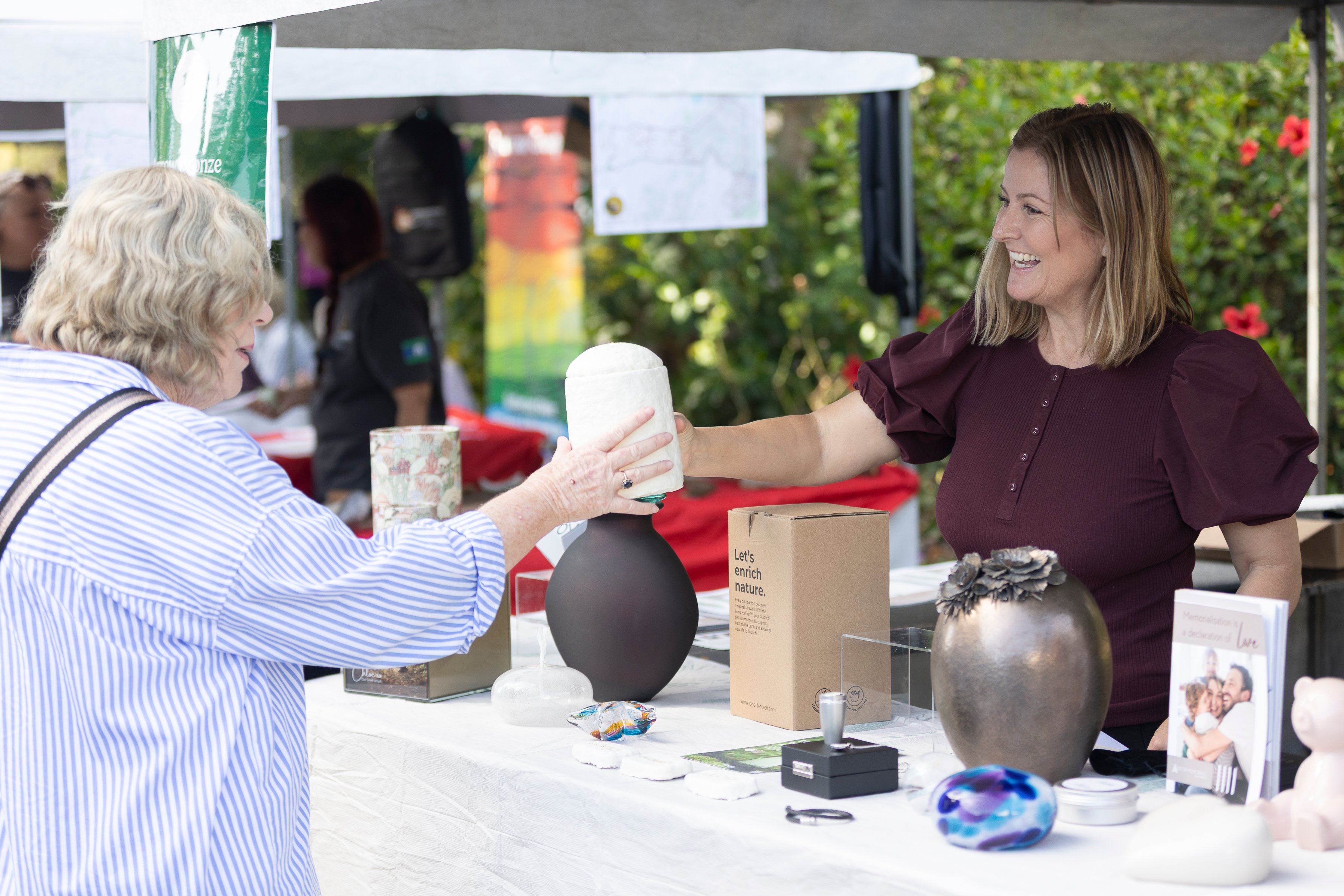 A woman in a burgundy shirt smiles while handing a ceramic piece to an older woman at an outdoor market stall displaying various ceramic items.