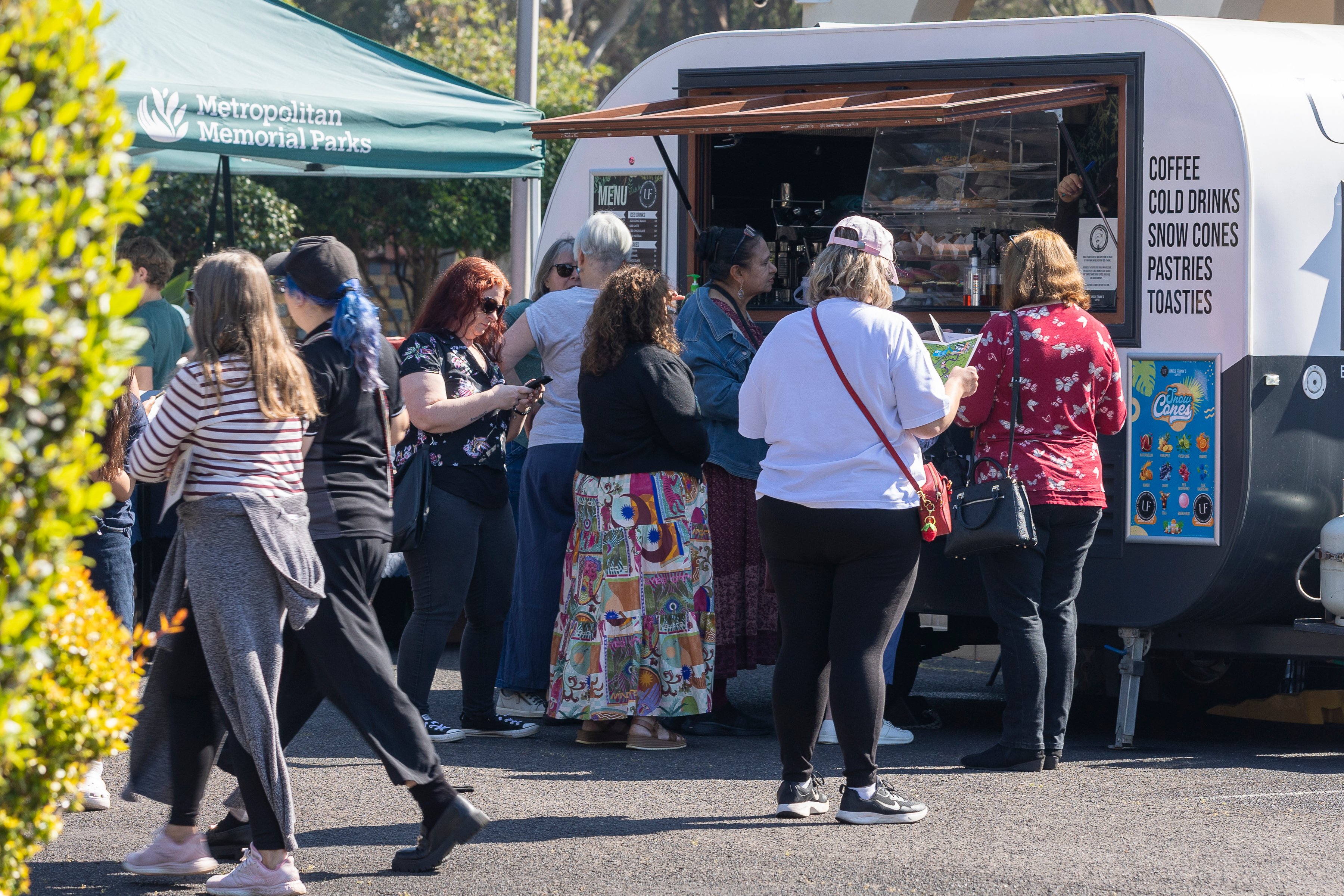 Visitors in colorful clothes queue at a coffee and pastry truck beneath a green park canopy, creating a lively, sunny outdoor scene.