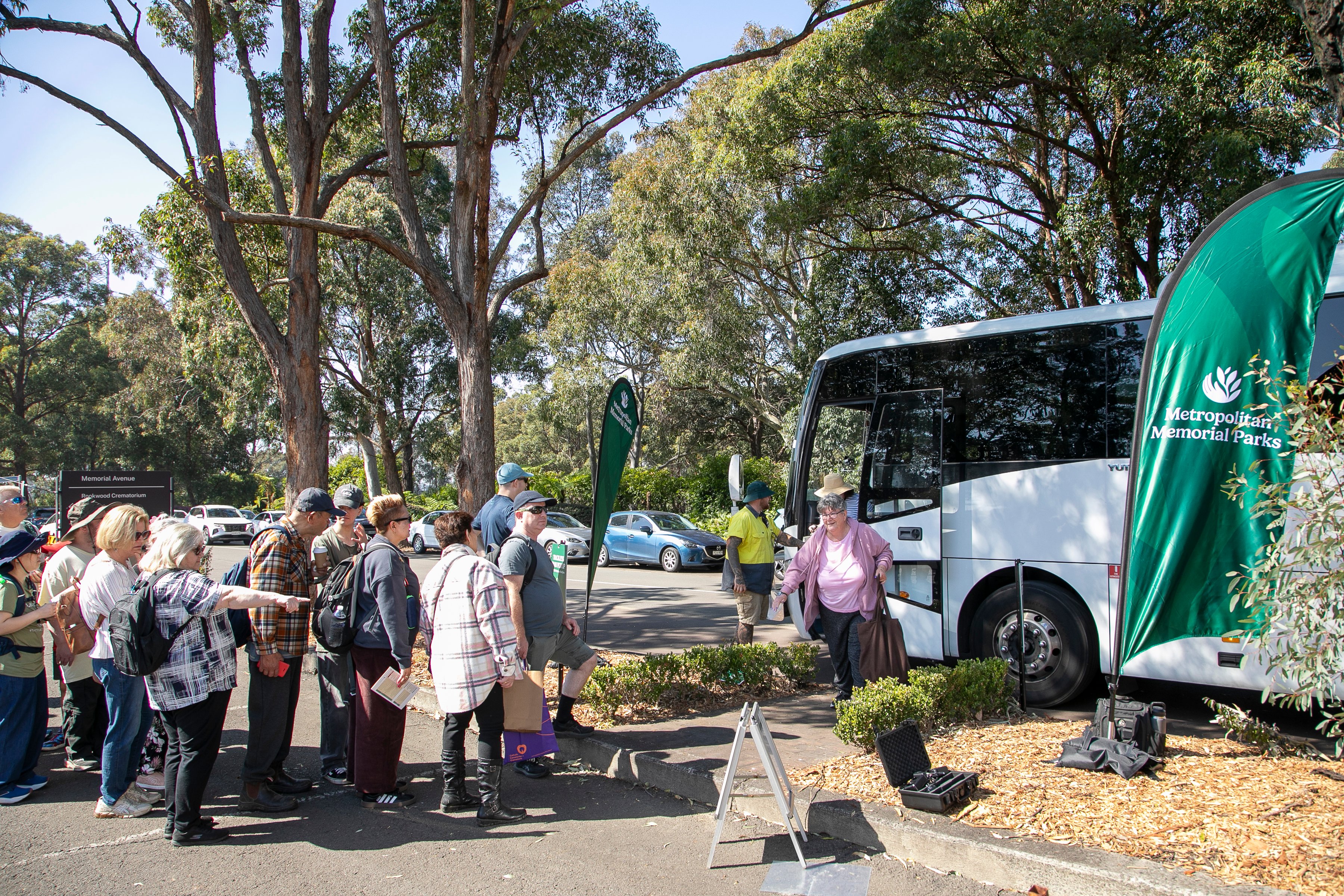 A group of adults gathers beside a white tour bus under shady trees, with event staff and green banners creating a calm community outing.