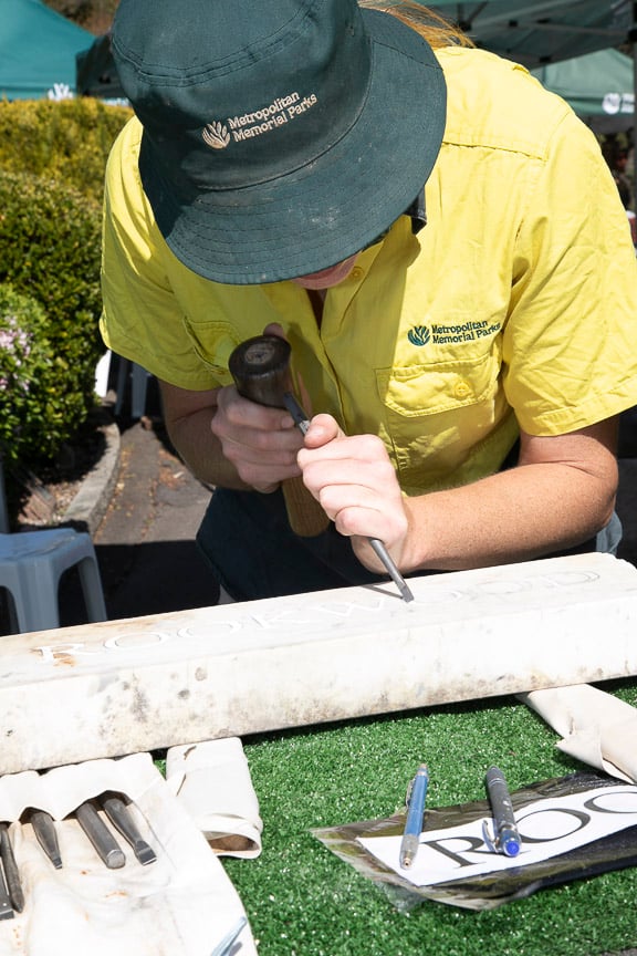 A Metroparks worker in a yellow shirt and wide-brimmed hat carefully chisels a mark into a white plank at an outdoor worksite.