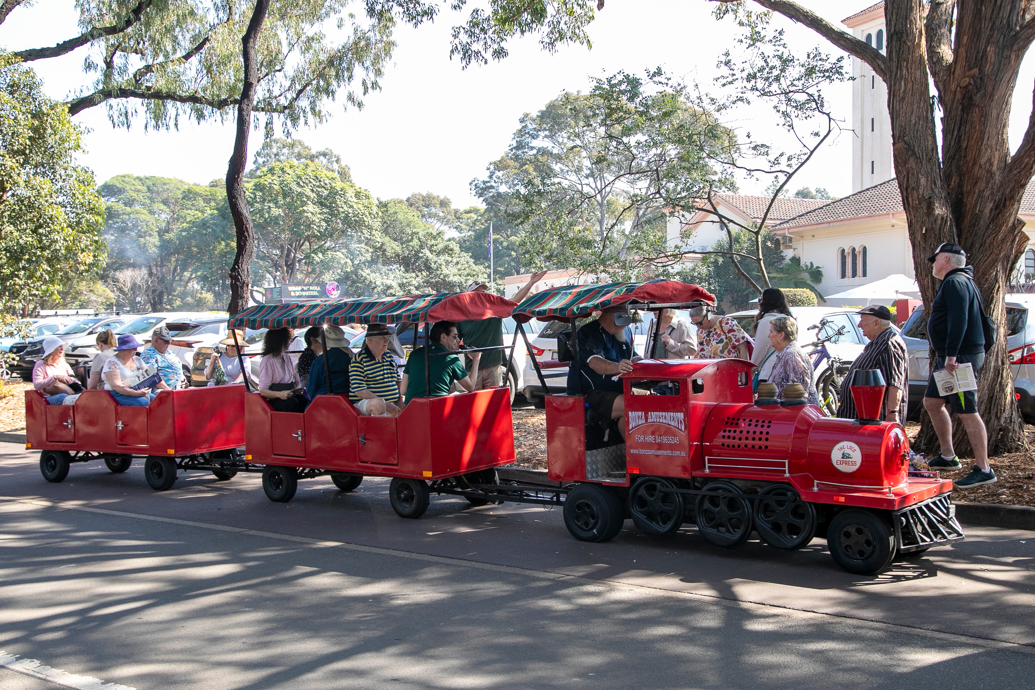 A bright red miniature train carries families through a sunny park, passing beneath large trees near a white building; the scene feels lively and relaxed.