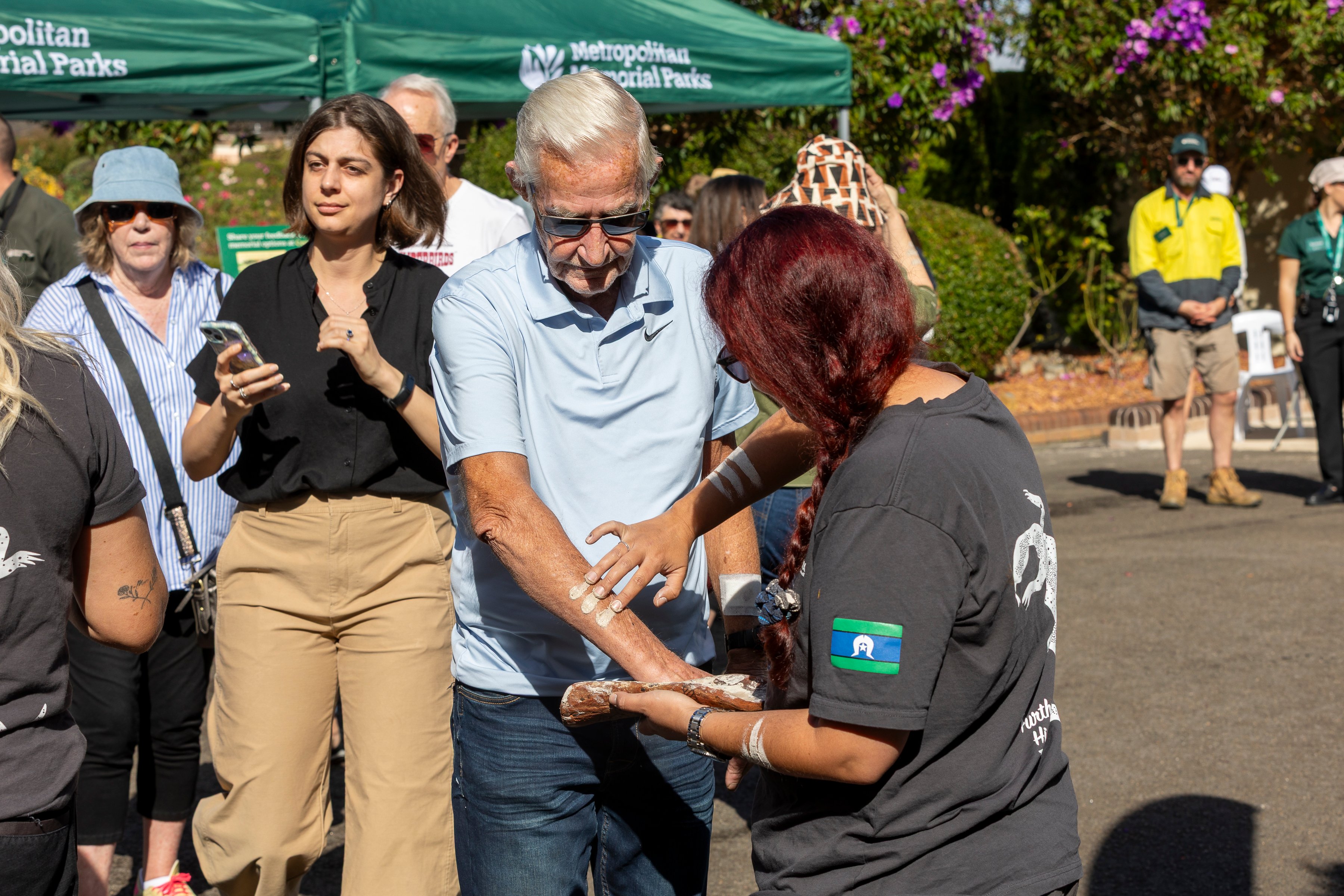 A woman with long hair applies white paint to an older man's arm during an outdoor cultural event. Onlookers stand around, with greenery in the background.