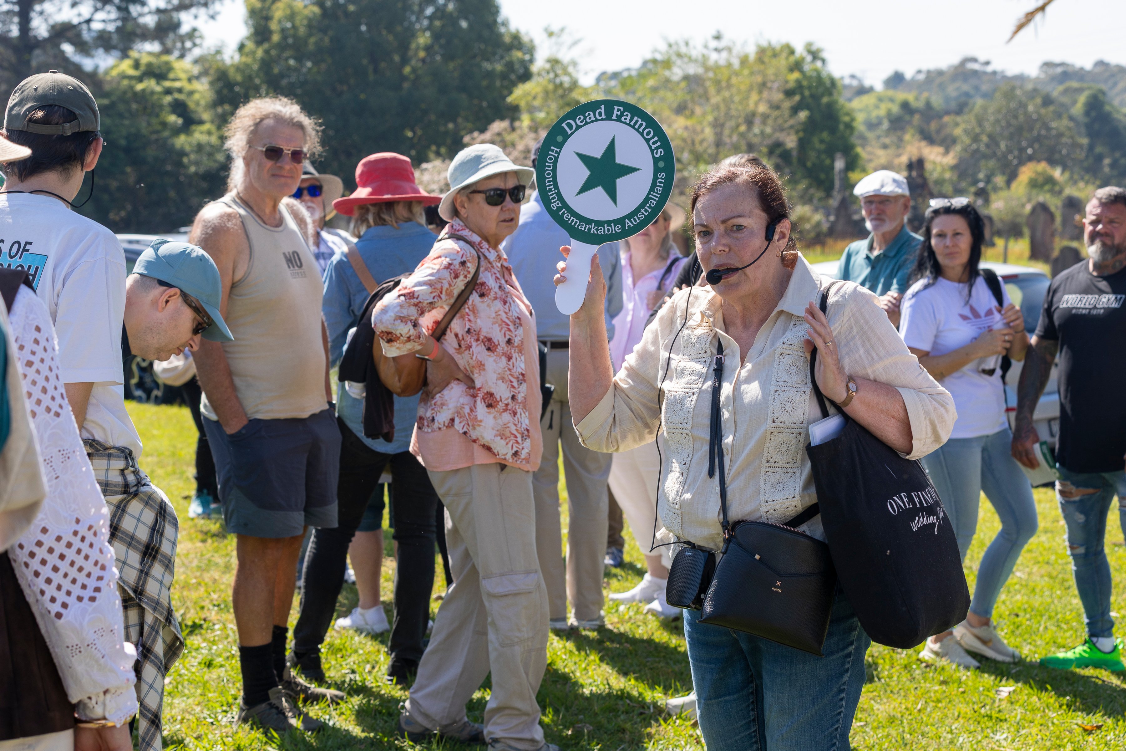 A woman holds a “Dead Famous” tour sign among a small group outdoors in a sunny park; visitors listen and chat in a relaxed, curious mood.