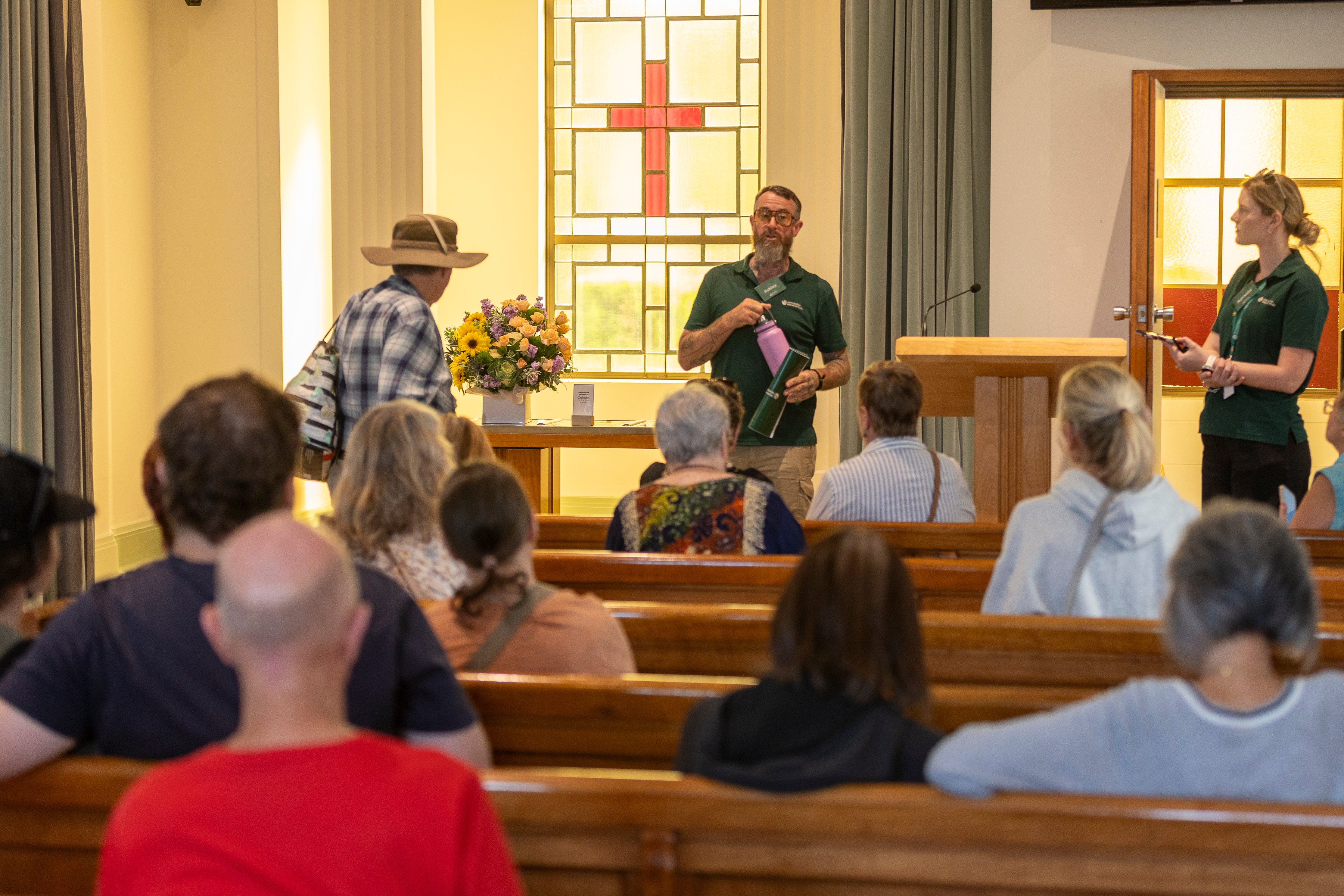 A group of people seated in a chapel listens to a speaker holding a pink item. A cross-shaped stained glass window and a flower arrangement are visible.