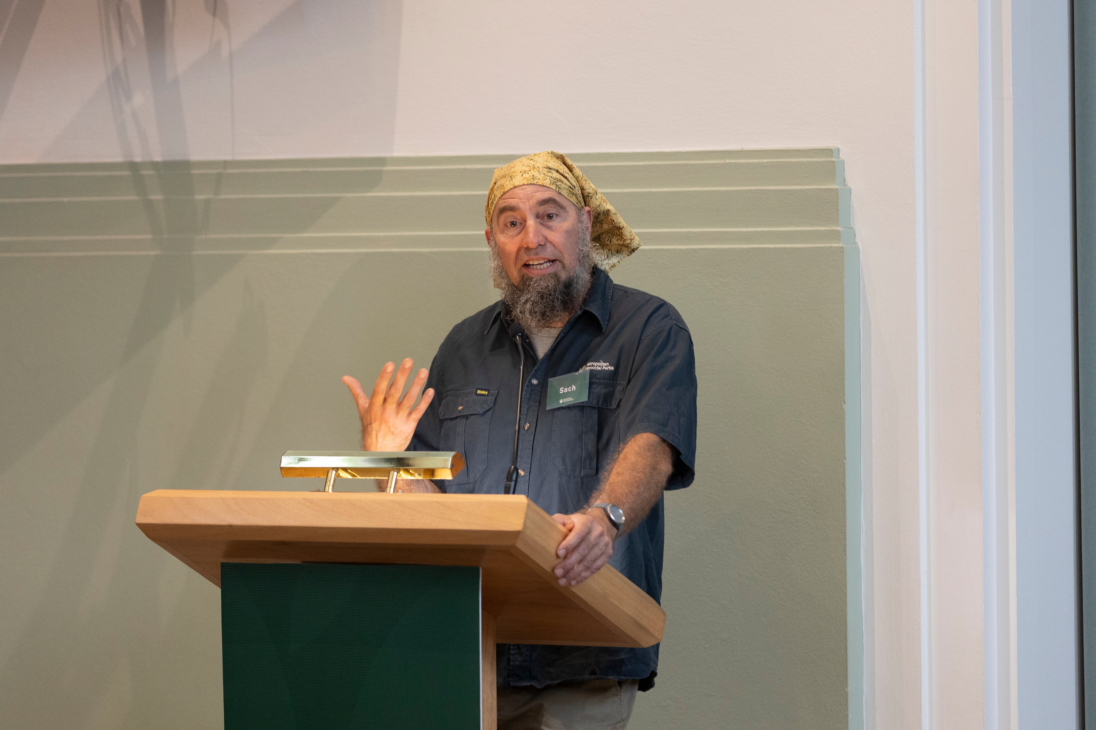 Man with a beard and headscarf speaks at a podium indoors. He gestures with one hand, wearing a navy shirt and name tag. Engaged expression, neutral-toned room.