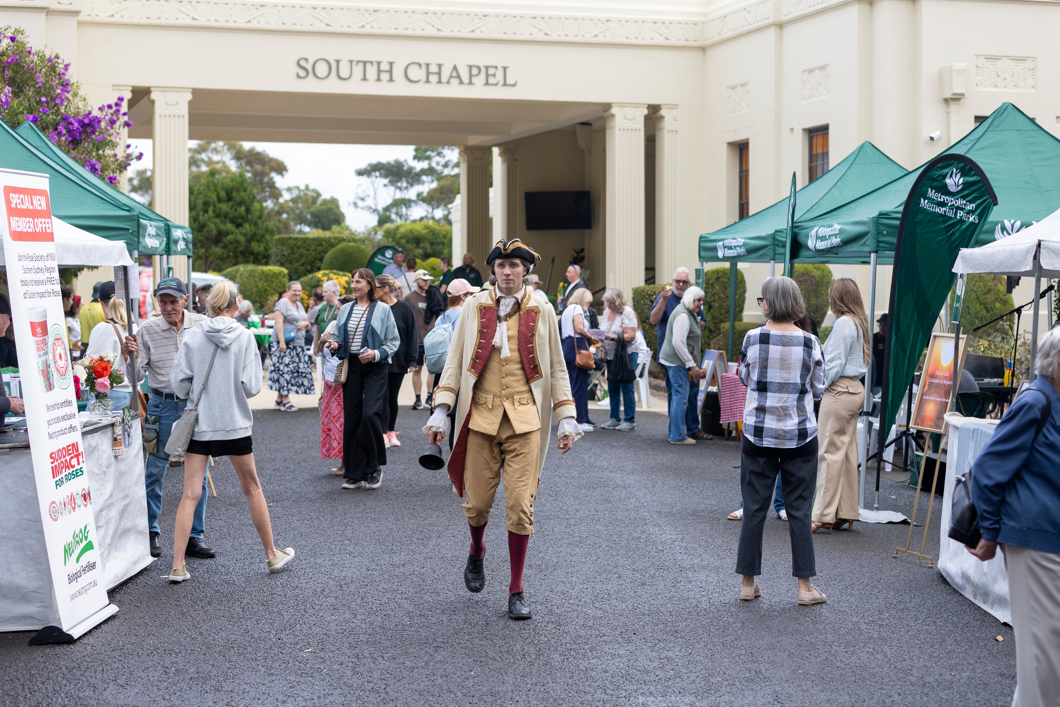A man in historical costume walks through a lively outdoor market with green tents, under a "South Chapel" sign. People are browsing stalls.