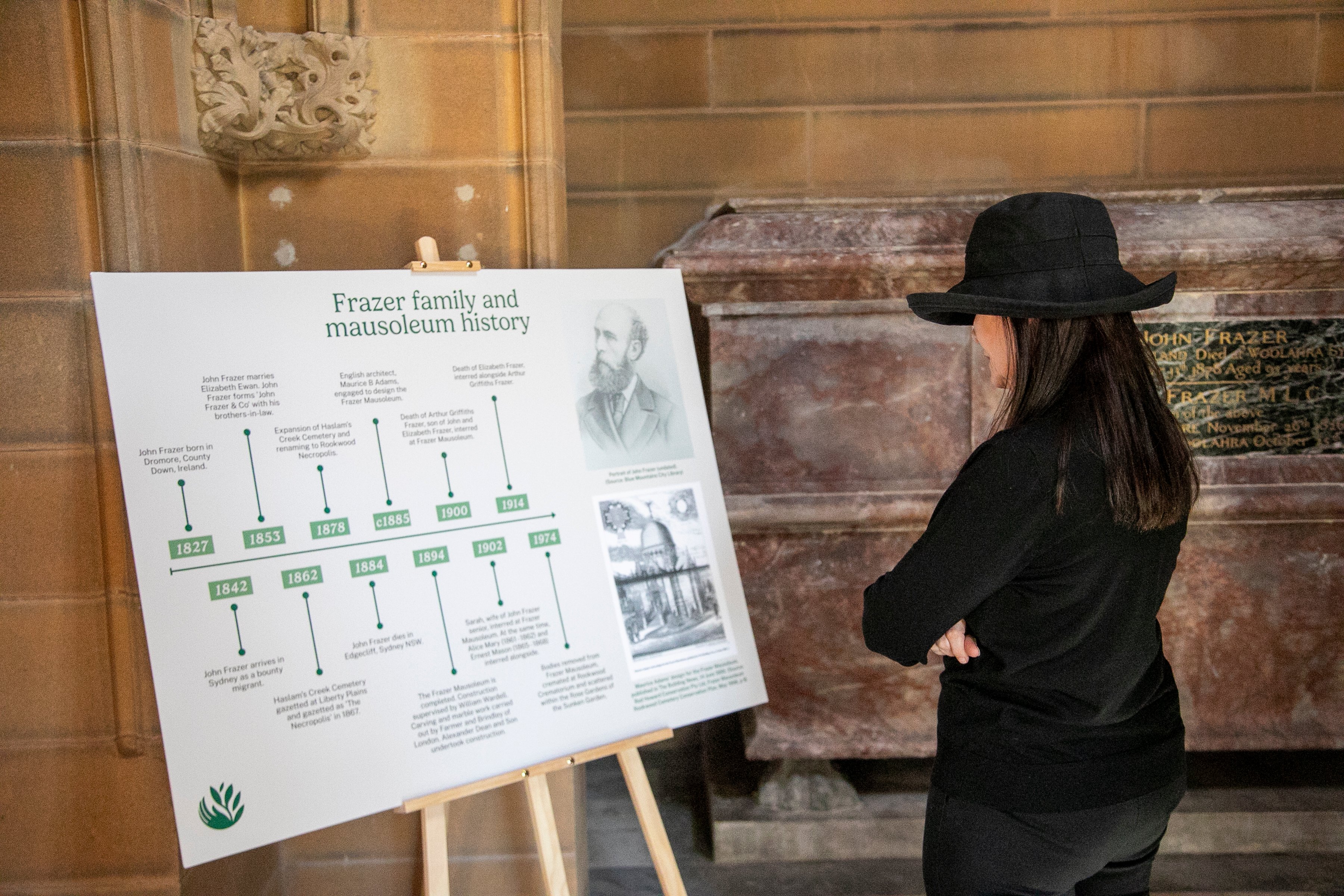 A visitor in a black hat studies a display tracing Frazer family and mausoleum history, standing thoughtfully beside a stone wall.