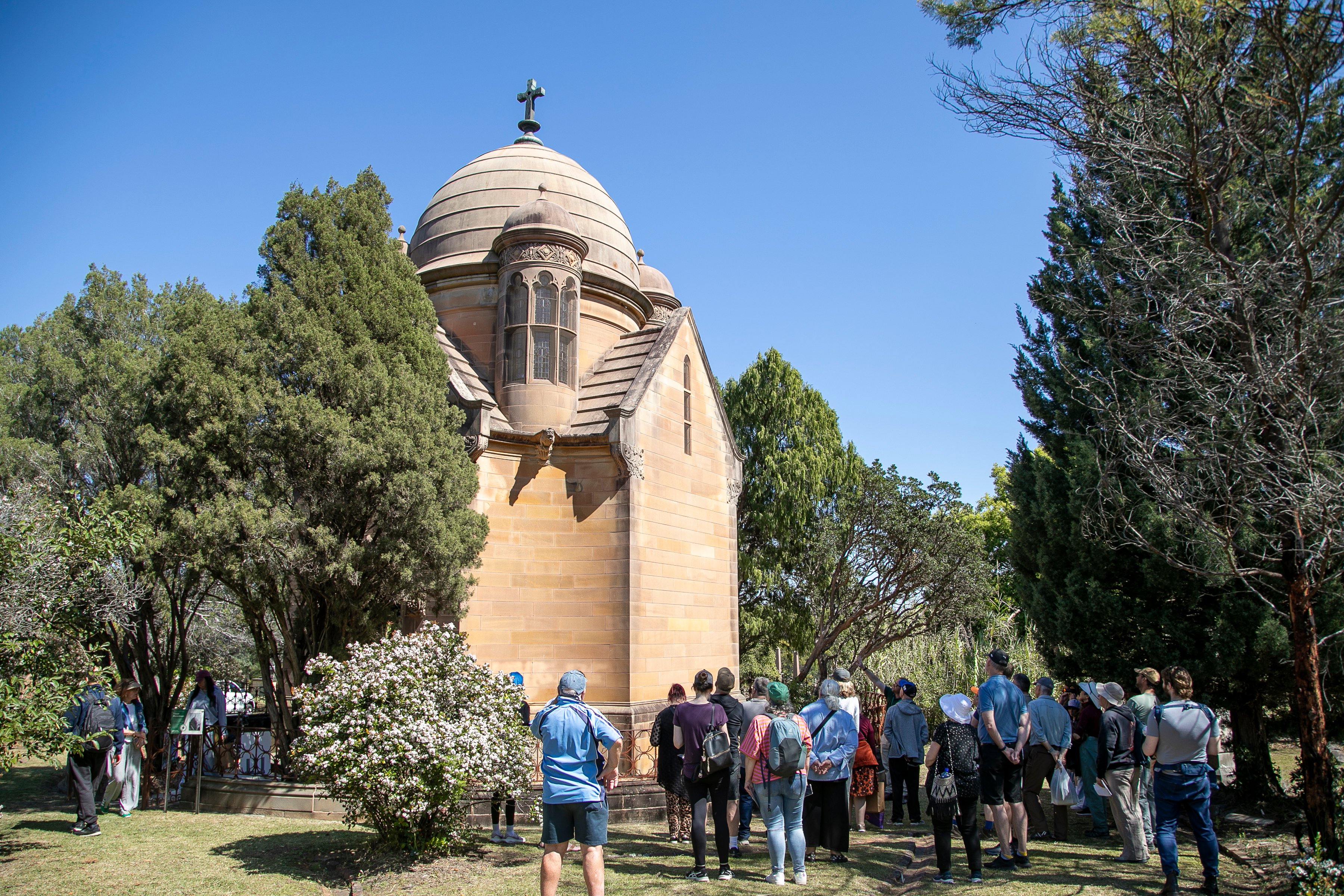 A tour group gathers beside a small historic stone chapel with a domed roof and cross, surrounded by trees under a clear blue sky.