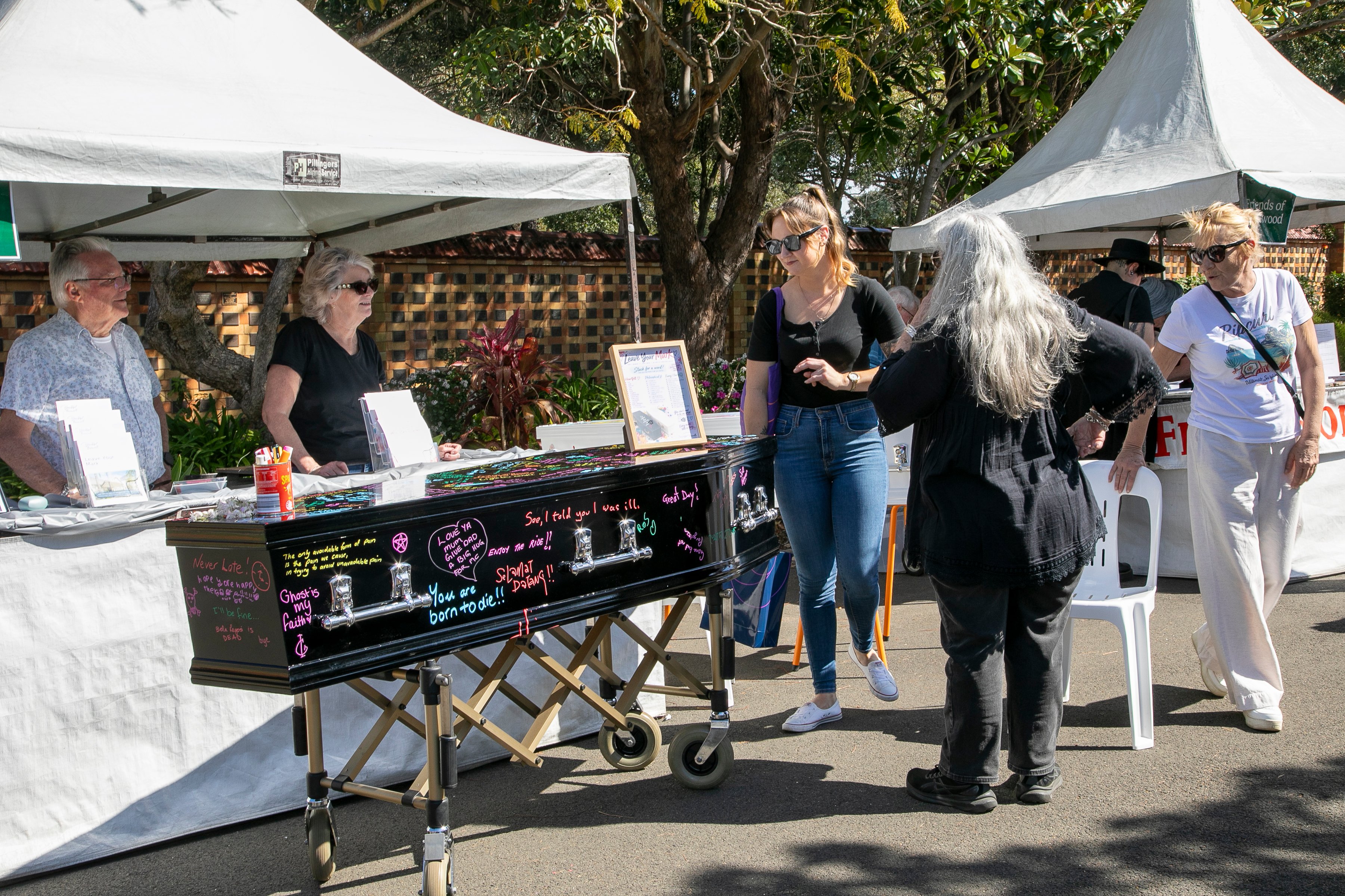 Shoppers browse a colorful handmade-goods stall beneath white market tents, chatting with vendors in bright sunlight; the scene feels lively and welcoming.