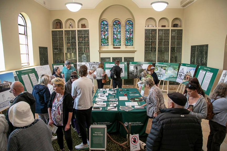 Visitors browse an environmental exhibition in a historic hall, examining green information boards and a central table; the atmosphere is lively and curious.