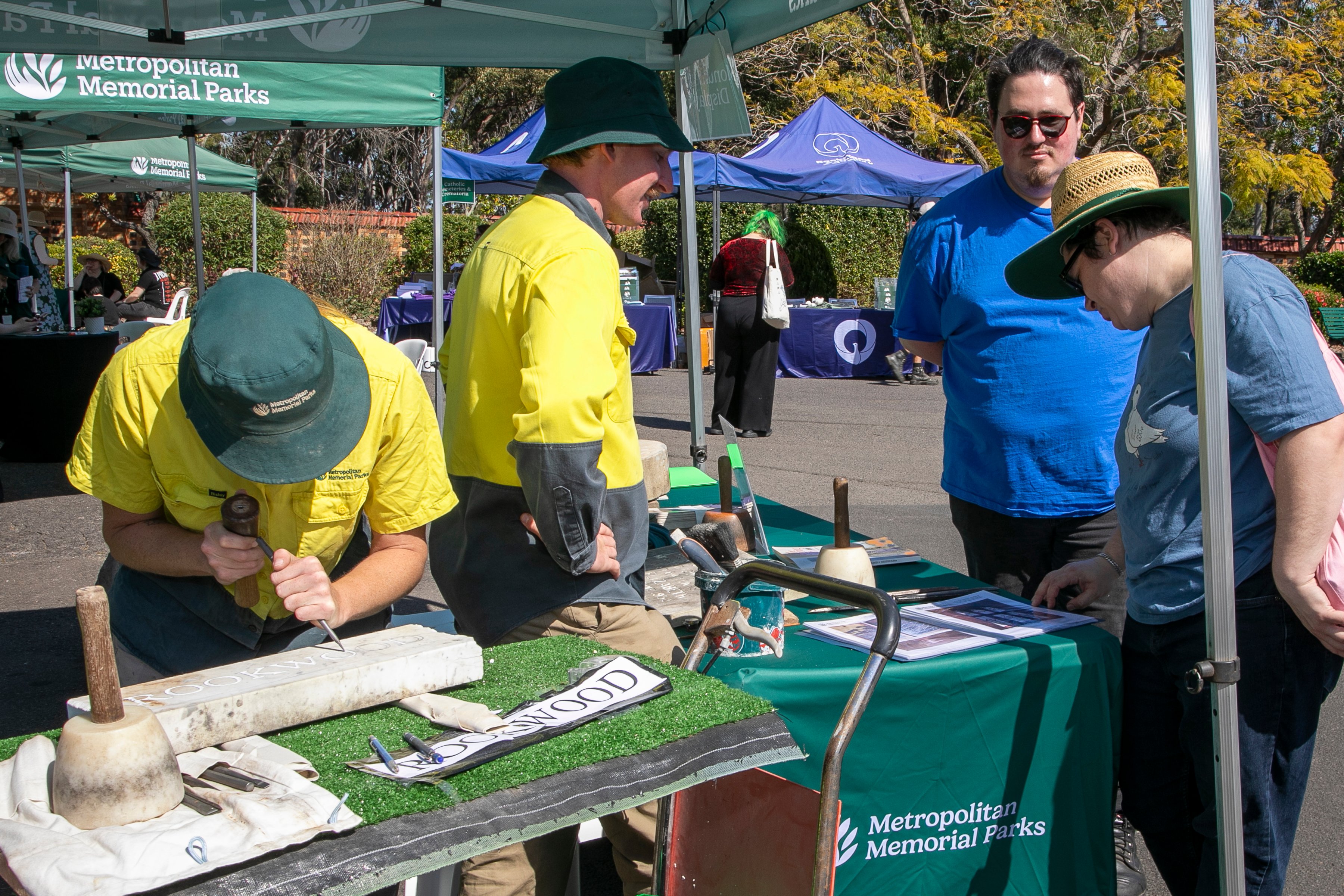 At an outdoor Metropolitan Memorial Parks booth, a yellow-shirted demonstrator works a chisel against a pale slab as visitors watch in the sunshine.