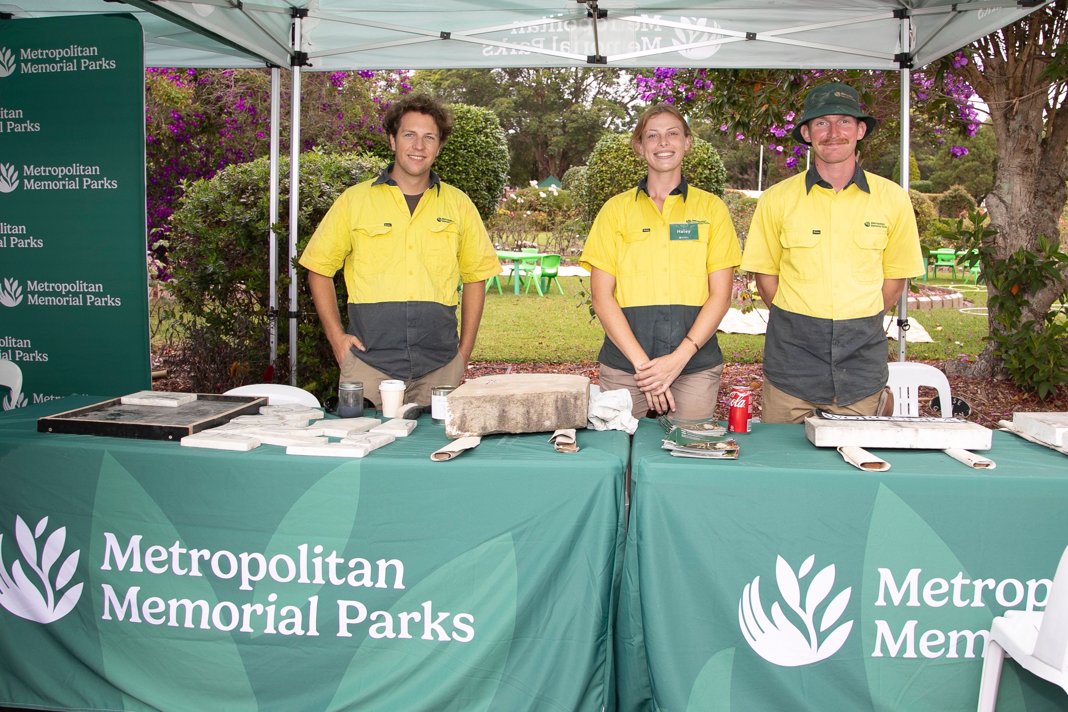 Three people in yellow uniforms stand behind a green booth with "Metropolitan Memorial Parks" branding. The setting is outdoors, surrounded by greenery and purple flowers, suggesting a welcoming and natural atmosphere.