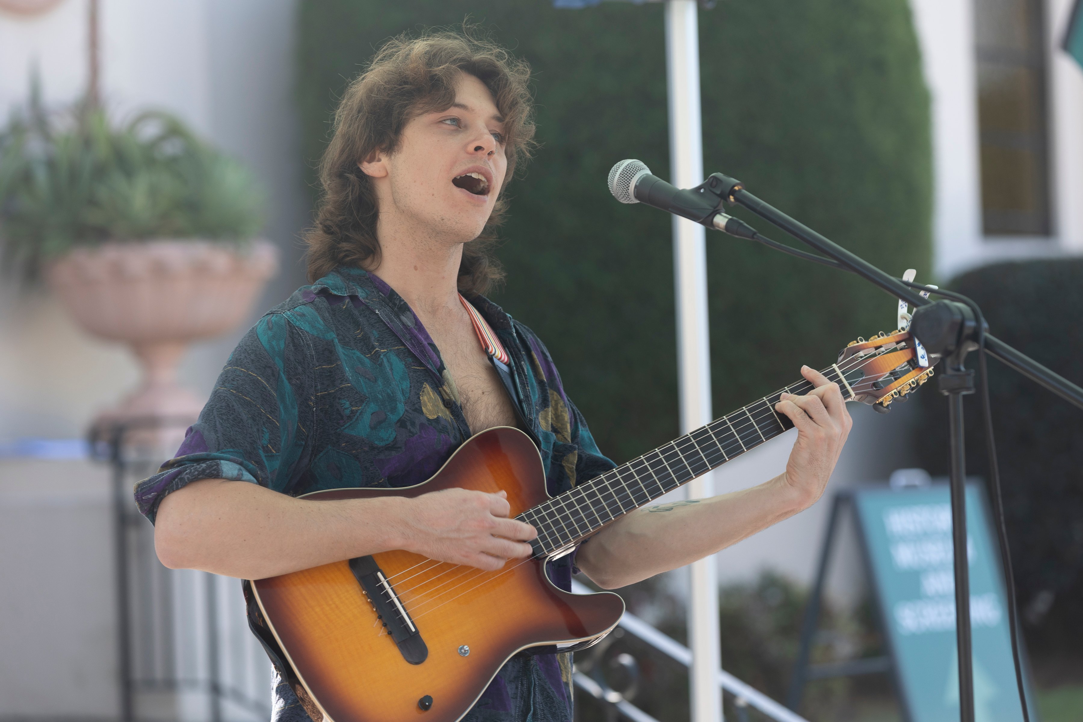 A young man sings passionately while strumming a sunburst acoustic guitar into a microphone outdoors, creating an energetic live-performance atmosphere.