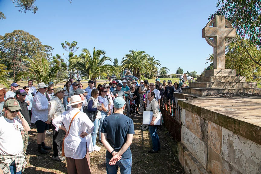 A tour group gathers beside an old stone cemetery monument topped with a Celtic cross, listening in bright sunshine amid palms and trees.